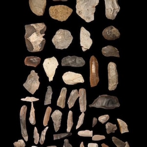 May include: A collection of various shaped, light-colored stones against a black background. The stones vary in size and shape, with some appearing to be chipped or flaked. The color palette includes shades of beige, gray, and brown, suggesting a natural, earthy aesthetic.