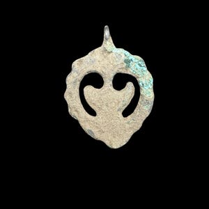 Può includere: Un ciondolo in metallo circolare e usurato con bordo smerlato e un intaglio centrale a forma di cuore o foglia stilizzata. La superficie ha un aspetto ruvido e invecchiato con macchie di ossidazione verde e un piccolo occhiello in alto.