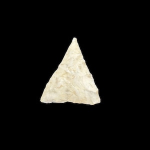 May include: A triangular stone arrowhead with a textured, off-white surface. The object features sharp, pointed edges and a flat base, displaying a chipped, knapped appearance with subtle variations in color and tone across its surface.