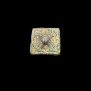 May include: A square-shaped bronze object with a weathered, greenish-brown patina. The surface features a raised central circular knob surrounded by four symmetrical, leaf-like embossed patterns that extend toward the corners of the square base.