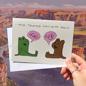 May include: A greeting card with the text "I MISS TALKING SHIT WITH YOU:(". The card features a green frog and a brown bear, each with a pink speech bubble containing scribbled lines. The card is held in a hand.