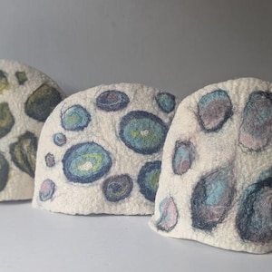 Peut inclure: Trois couvre-théières en laine feutrée en forme de dôme de couleur blanc cassé. Chaque pièce présente un motif unique de formes circulaires et ovales dans des tons vert olive, bleu foncé, bleu clair et rose pâle, créant un aspect texturé.