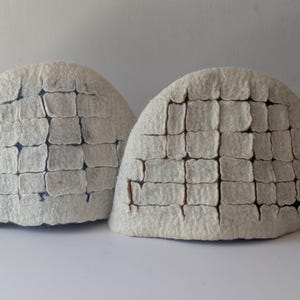 May include: Two dome-shaped, off-white felt sculptures, resembling igloos. Each has a textured surface with a grid-like pattern of rectangular blocks. The background is a plain, light-colored surface.