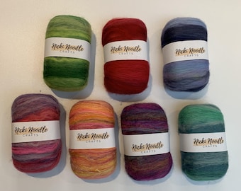 May include: Seven skeins of yarn arranged in two rows, each wrapped in a white paper band labeled Nicki Noodle CRAFTS. The yarn colors include solid green, solid red, blue gradient, pink and purple gradient, orange and yellow, purple, and teal.