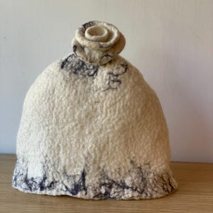 May include: A cream-colored felt hat with a decorative rose on top. The hat has dark grey accents around the base and on the crown. The hat is sitting on a wooden surface.