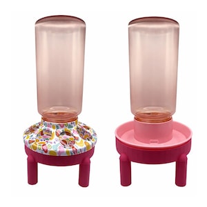 May include: Two pet feeders. One has a pink base with a patterned top featuring fruits and other designs. The other has a solid pink base. Both have a clear, cylindrical water bottle on top. The feeders are elevated on short legs.