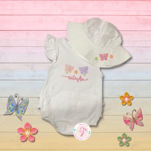 Pode incluir: Conjunto de bebé branco com macacão e chapéu de sol. O macacão apresenta bordados de borboletas e flores com o nome "natasha". O chapéu de sol tem bordados a condizer de borboletas e flores. Decorações adicionais de borboletas e flores.