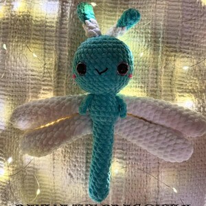 Puede incluir: Un peluche de libélula de ganchillo turquesa y blanco, con ojos grandes y oscuros y mejillas rosadas. La libélula tiene alas blancas y un cuerpo largo turquesa. El texto "DAHLIA THE DRAGONFLY" está en la parte inferior de la imagen.