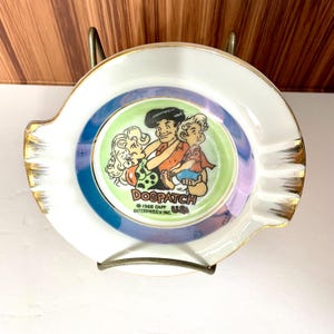 May include: White ceramic ashtray with a gold rim and a colorful cartoon image of Dogpatch characters. The center features a green circle with the text "DOGPATCH" and "©1968 CAPP ENTERPRISES: INC. USA". The ashtray has a curved shape with a small notch.