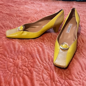 Op de afbeelding: Een paar gele leren pumps met een vierkante neus en een kleine hak. De neus en de hielkap hebben een slangenprint. Elke schoen heeft een zilverkleurige gesp.