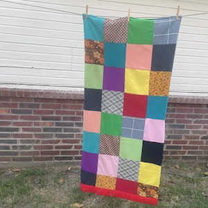 May include: A colorful patchwork quilt hanging on a clothesline. The quilt is made of various squares in different colors and patterns, including polka dots, plaid, and floral designs. The quilt has a red border.
