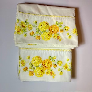 May include: Folded white bed sheets with a yellow floral pattern featuring roses and other flowers. The edges are trimmed with yellow scalloped detailing. The floral design is printed across the bottom of the sheets.