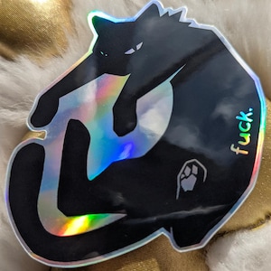 May include: A black cat sticker with a holographic finish, featuring the word "fuck." The cat is in a playful pose, with a paw print detail. The sticker is set against a backdrop of white fur and gold fabric.