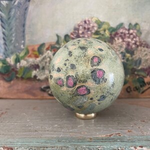 May include: A decorative sphere with a speckled green and pink pattern, mounted on a gold-coloured base. The sphere is a focal point, set against a blurred background with floral elements and a vintage aesthetic.
