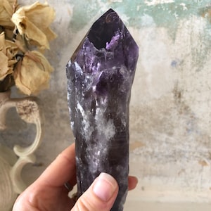 Può includere: Una grande punta di cristallo di ametista viola con una texture naturale e ruvida. Il cristallo è appuntito in cima e ha una base piatta.