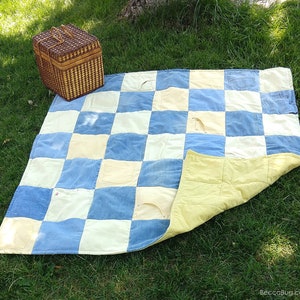 Op de afbeelding: Een handgemaakte picknickdeken gemaakt van gerecyclede denim en crèmekleurige stoffen vierkanten. De deken heeft een dambordpatroon met denim zakken en een gele, bedrukte onderkant. Een bruine picknickmand van riet staat ernaast op het gras.