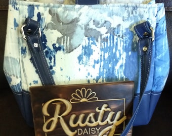 Może przedstawiać: Torba typu tote w kolorze niebieskim i białym z niebieskimi skórzanymi wykończeniami i uchwytami. Torba leży na drewnianym znaku z napisem "Rusty Daisy" złotymi literami.