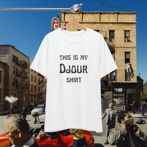 May include: A white short-sleeved t-shirt featuring the black printed text THIS IS MY Djour SHIRT centered on the front. The garment has a classic crew neckline and a straight hem, presented on a wooden hanger against a city street backdrop.
