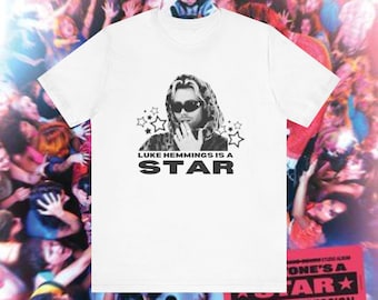 May include: White t-shirt featuring a black and white image of a person wearing sunglasses, surrounded by stars. The text "LUKE HEMMINGS IS A STAR" is printed below the image in black. The background shows a crowd of people.