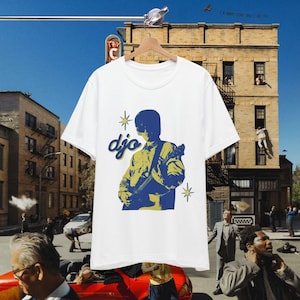 May include: White t-shirt with a blue and yellow illustration of a guitarist, the word "djo" in a stylized font, and star accents. The t-shirt is displayed on a wooden hanger.