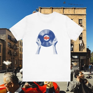 May include: White t-shirt with a blue and red graphic of a vinyl record held by hands. The word "djo" is printed on the record. A casual wear item.