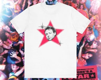 May include: White t-shirt featuring a red star graphic with a man's face in the center. The shirt has a crew neck and short sleeves. The background shows a crowd of people.