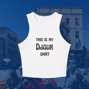 May include: A white sleeveless crop top featuring a round neckline and black text printed on the front that reads THIS IS MY Djour SHIRT. The garment has a racerback style cut with wide shoulder straps and a cropped hemline that sits at the waist.