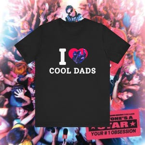 May include: A black short-sleeved t-shirt featuring the white text I and COOL DADS with a red heart graphic in the center. Inside the heart is a small photographic image of a person wearing sunglasses. The shirt has a crew neckline.