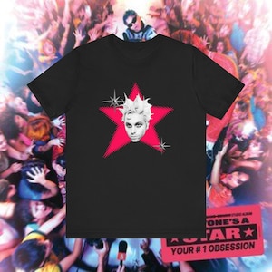 Puede incluir: Camiseta negra con un gráfico de una cara con pelo blanco dentro de una estrella rosa. La estrella tiene un patrón de puntos y está rodeada de explosiones de estrellas. La camiseta forma parte de una colección temática musical.