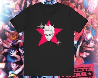May include: Black t-shirt featuring a graphic of a face with white hair inside a pink star. The star has a dotted pattern and is surrounded by starbursts. The shirt is part of a music-themed collection.