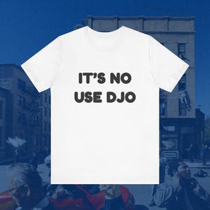 Könnte beinhalten: Weißes T-Shirt mit dem Text "IT'S NO USE DJO" in fetter, schwarzer Schrift. Das T-Shirt hat einen Rundhalsausschnitt. Der Hintergrund zeigt eine verschwommene urbane Szene, was auf eine Außenumgebung hindeutet.