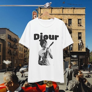 May include: A white short-sleeved t-shirt featuring a black graphic print of a person playing an electric guitar. Above the graphic, the word "Djour" is printed in a bold, rounded black font. The shirt is displayed on a wooden hanger.