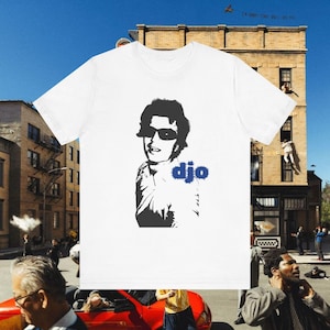 May include: White t-shirt with a black and white graphic of a person wearing sunglasses, and the word "djo" in blue. The t-shirt has a crew neck.