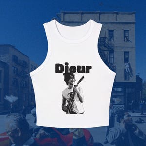 May include: A white sleeveless crop top featuring a black graphic print of a person playing an electric guitar. Above the figure, the word "Djour" is printed in a bold, rounded black font. The garment has a high, rounded neckline.