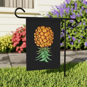 May include: A black garden flag featuring a detailed illustration of a pineapple. The pineapple is depicted in shades of yellow and orange, with green leaves at the base. The flag is displayed on a black metal stand in a garden setting.