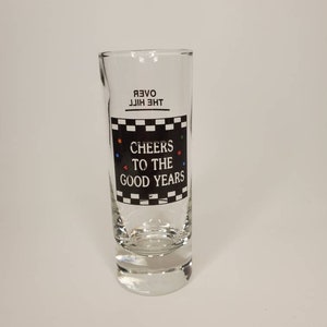 May include: A clear glass shot glass with a black and white checkered design and the text "Cheers to the Good Years" printed on the side. The text "Over the Hill" is printed upside down at the top of the glass.