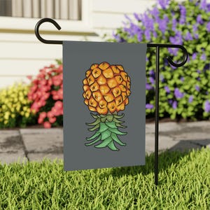 May include: A gray garden flag featuring a cartoon pineapple design. The pineapple is depicted in shades of orange and yellow, with green leaves at the base. The flag is displayed on a black metal stand in a grassy yard.