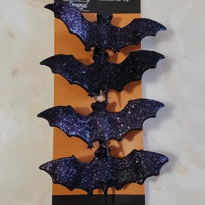 May include: Four black glitter bat clips with a Halloween Bat Clip label. The label has a black cat and pumpkin graphic and the text "Seasonal Inc."