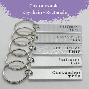 May include: Five rectangular silver-toned metal keychains, each attached to a circular split ring. The metal tags feature various stamped fonts and styles, with the text "CUSTOMIZE THIS" or "Customize This" imprinted on each individual piece.