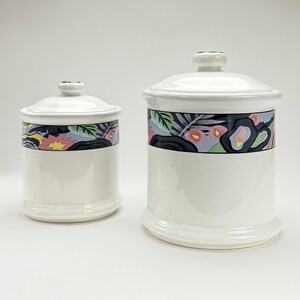 May include: Two white ceramic storage jars with lids. Each jar has a decorative band with a floral and leaf design in pink, black, and green. The jars are different sizes, with a smaller one to the left and a larger one to the right.