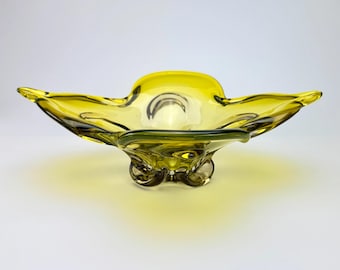 May include: A vintage, yellow-green glass bowl with an unusual, asymmetrical design. The bowl has a wide, open shape with flared edges and rests on three small, rounded feet. The glass has a glossy finish, and the bowl is likely used for decorative purposes.