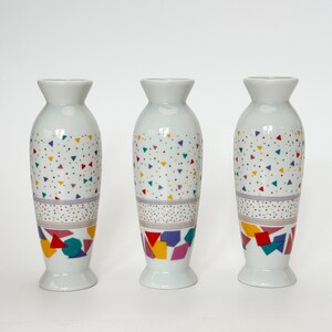 May include: Three white ceramic vases with a colourful geometric design. The vases feature a pattern of multicoloured triangles and dots on the upper portion, with a band of dots and lines. The lower section has a design of colourful geometric shapes.