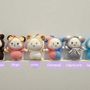 May include: Seven crocheted plush toys representing the zodiac signs. The toys are brown, pink, white, pink and blue, gray, and blue. The signs are Sagittarius, Virgo, Aries, Gemini, Capricorn, and Aquarius.