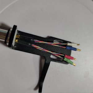 May include: A black and silver audio cartridge with multi-colored wires and gold-tipped connectors. The wires are red, green, blue, and white, and are twisted together. The cartridge is on a white surface.