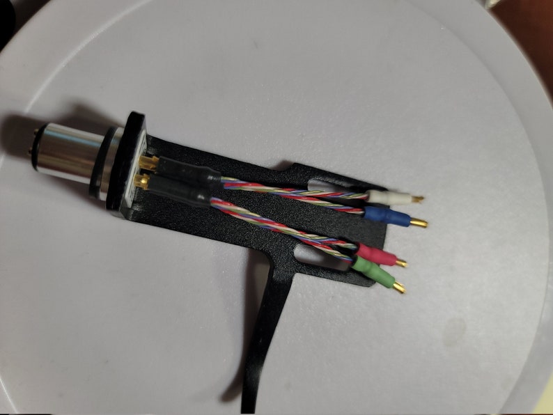 May include: A black and silver audio cartridge with multi-colored wires and gold-tipped connectors. The wires are red, green, blue, and white, and are twisted together. The cartridge is on a white surface.