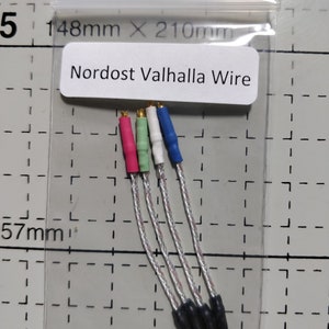 May include: A clear plastic bag containing four Nordost Valhalla wires. Each wire has a different colored connector: pink, green, white, and blue. The wires are silver and black. The text "Nordost Valhalla Wire" is printed on a white label.