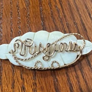 May include: A white mother of pearl leaf-shaped brooch with a gold wire inscription that reads "Priscilla".