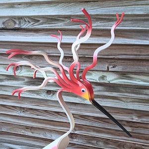 May include: A whimsical metal sculpture of a bird with a long, black beak and a vibrant red, yellow, and white color scheme. The bird's head features a crest of flowing, curved elements. It stands on a black base, creating a striking decorative piece.