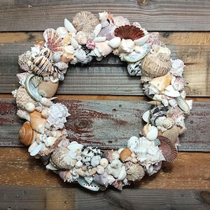 May include: A decorative wreath composed of various seashells in shades of white, brown, and tan. The shells are arranged in a circular shape, creating a coastal or nautical theme. The wreath is displayed against a rustic wooden background.