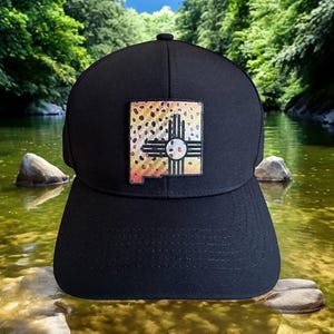 May include: Black baseball cap with a square patch featuring a New Mexico state outline and a fish pattern. The patch has a brown, tan, and orange speckled design with a white and red sun symbol. The cap is set against a scenic river background.